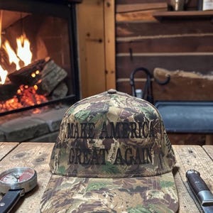 Puede incluir: Gorra de b&eacute;isbol de camuflaje con el texto "MAKE AMERICA GREAT AGAIN" en negro. La gorra est&aacute; sobre una mesa de madera con una br&uacute;jula y un cuchillo. Una chimenea con le&ntilde;os ardiendo se encuentra en el fondo.