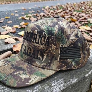 Puede incluir: Gorra de b&eacute;isbol de camuflaje con el texto "AMERICA AGAIN" en letras negras. La gorra presenta un peque&ntilde;o parche de la bandera estadounidense. El sombrero est&aacute; hecho de un patr&oacute;n de camuflaje con tonos de verde, marr&oacute;n y negro.