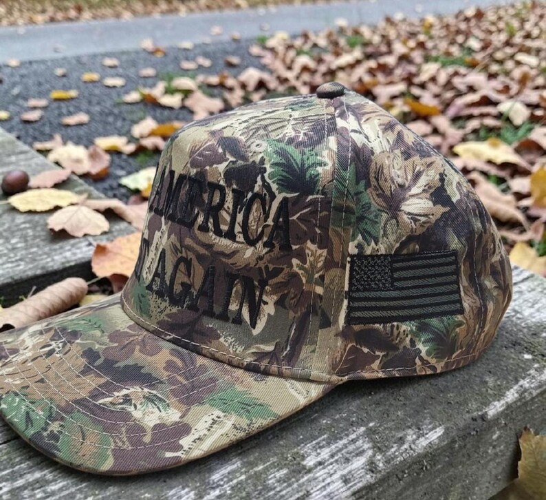 Puede incluir: Gorra de b&eacute;isbol de camuflaje con el texto "AMERICA AGAIN" en letras negras. La gorra presenta un peque&ntilde;o parche de la bandera estadounidense. El sombrero est&aacute; hecho de un patr&oacute;n de camuflaje con tonos de verde, marr&oacute;n y negro.