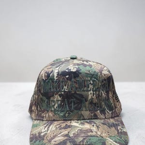 May include: Camouflage baseball cap with the embroidered text "MAKE AMERICA GREAT AGAIN" in green. The cap features a curved brim and a button on top. The fabric has a pattern of green, brown, and tan.