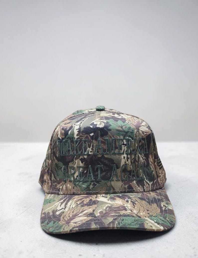 May include: Camouflage baseball cap with the embroidered text "MAKE AMERICA GREAT AGAIN" in green. The cap features a curved brim and a button on top. The fabric has a pattern of green, brown, and tan.