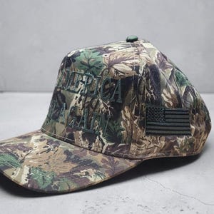 May include: Camouflage baseball cap with the embroidered text "AMERICA AGAIN" in green. Features a small American flag patch on the side. The cap has a curved brim and a button on top.