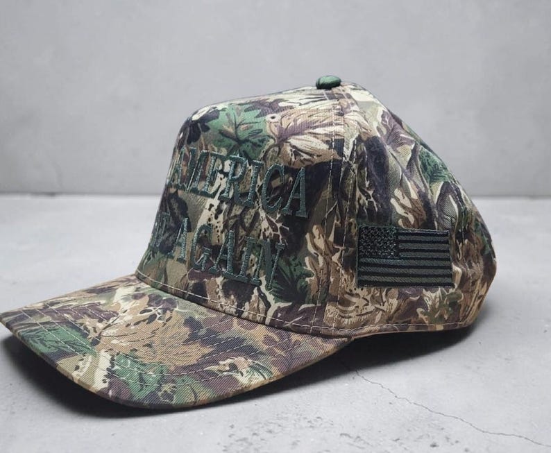 May include: Camouflage baseball cap with the embroidered text "AMERICA AGAIN" in green. Features a small American flag patch on the side. The cap has a curved brim and a button on top.
