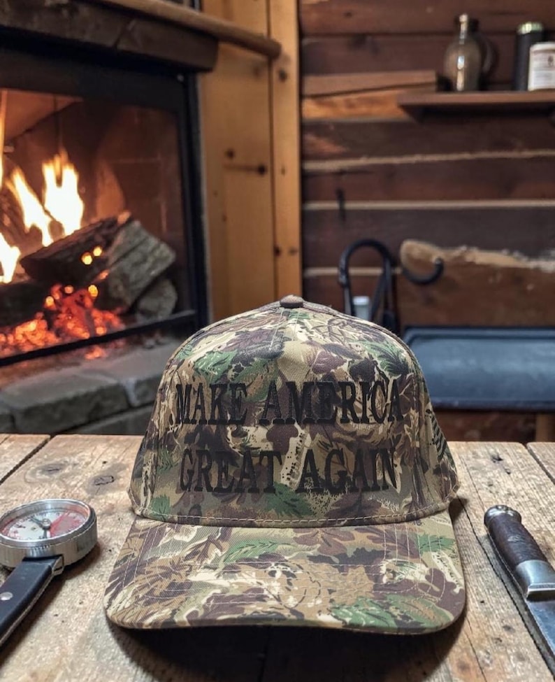 Puede incluir: Gorra de b&eacute;isbol de camuflaje con el texto "MAKE AMERICA GREAT AGAIN" en negro. La gorra est&aacute; sobre una mesa de madera con una br&uacute;jula y un cuchillo. Una chimenea con le&ntilde;os ardiendo se encuentra en el fondo.