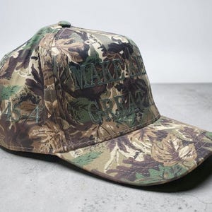 May include: Camouflage baseball cap with the embroidered text "MAKE AMERICA GREAT" in green. The cap features a floral pattern in shades of green, brown, and tan. The number "45-45" is embroidered on the side of the cap.