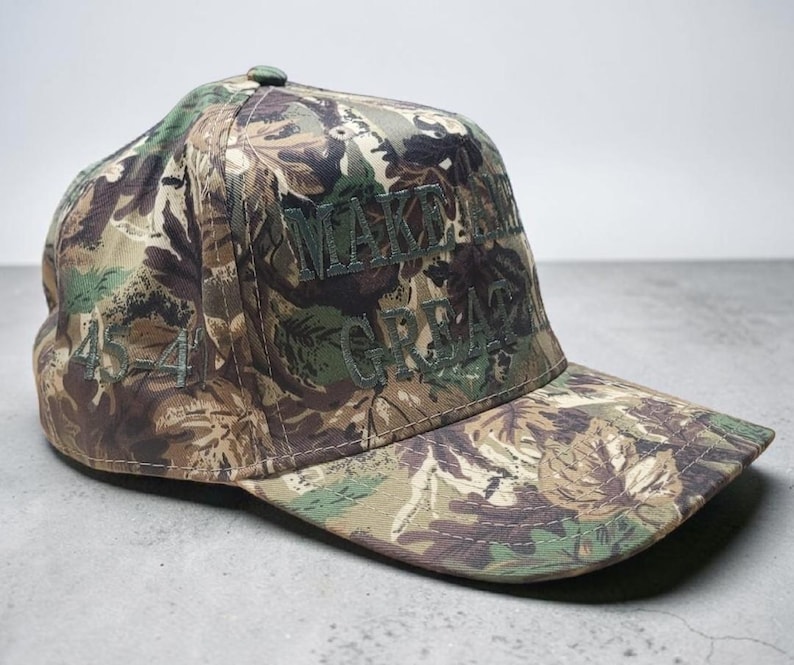 May include: Camouflage baseball cap with the embroidered text "MAKE AMERICA GREAT" in green. The cap features a floral pattern in shades of green, brown, and tan. The number "45-45" is embroidered on the side of the cap.