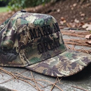 Puede incluir: Gorra de b&eacute;isbol de camuflaje con las palabras "MAKE AMERICA GREAT" bordadas en negro. La gorra presenta una visera curva y un bot&oacute;n en la parte superior. La gorra est&aacute; hecha de una tela con un patr&oacute;n de verde, marr&oacute;n y beige.