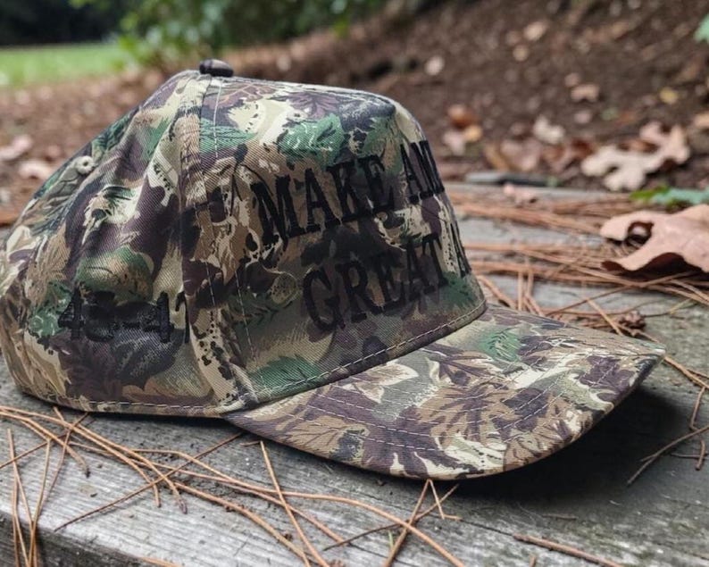 Puede incluir: Gorra de b&eacute;isbol de camuflaje con las palabras "MAKE AMERICA GREAT" bordadas en negro. La gorra presenta una visera curva y un bot&oacute;n en la parte superior. La gorra est&aacute; hecha de una tela con un patr&oacute;n de verde, marr&oacute;n y beige.