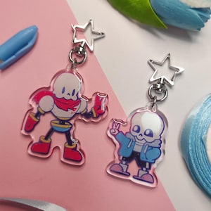 Può includere: Due portachiavi in ​​acrilico trasparente con personaggi dei cartoni animati del videogioco Undertale. Il portachiavi a sinistra presenta Papyrus, uno scheletro alto che indossa una sciarpa rossa e pantaloncini blu. Il portachiavi a destra presenta Sans, uno scheletro più basso che indossa una giacca blu e pantaloncini blu. Entrambi i portachiavi hanno un ciondolo a forma di stella argentata.