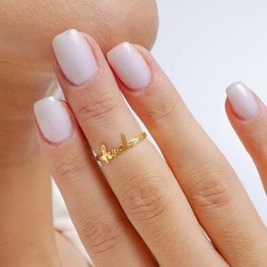 May include: A gold ring with the word "huda" written on it. The ring is worn on the middle finger of a person's hand.