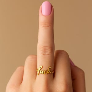 May include: A gold-colored ring with the word "fuck" in cursive script. The ring is on a finger with a pink nail polish. The background is a solid tan color.