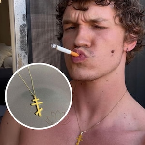 May include: A gold-colored Orthodox cross pendant necklace. The pendant features a three-barred cross design, suspended from a delicate chain. The necklace is displayed against a neutral background, highlighting the jewelry's details.