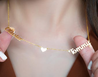 May include: A gold-colored necklace with the names "Olivia" and "Tommy" in cursive script. A small heart charm is between the names. The necklace is being held up by a person's fingers.