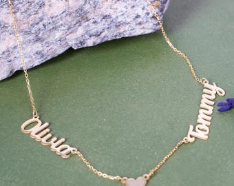 May include: A gold-colored necklace with the names "Olivia" and "Tommy" in cursive script. A small heart charm connects the two names. The necklace is displayed on a green surface with a rock and purple flowers in the background.