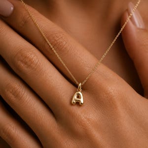 Könnte beinhalten: Eine goldfarbene Halskette mit einem kleinen, aufgeblähten Buchstaben-A-Anhänger, der an einer zarten, dünnen Kette hängt. Der Anhänger hat eine glatte, polierte Oberfläche mit einer abgerundeten, dreidimensionalen Form, die das Licht einfängt.