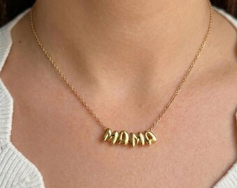 May include: A gold necklace featuring the word "MAMA" in individual, rounded letters. The necklace is suspended from a delicate chain and worn around the neck. The background is a light-coloured top.