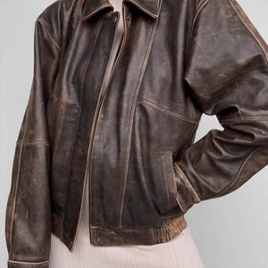 May include: A brown leather jacket with a distressed, vintage appearance. The jacket has a collared neckline, a zip closure, and a pocket on the left side. The jacket has a ribbed hem and cuffs. The leather has a worn appearance.