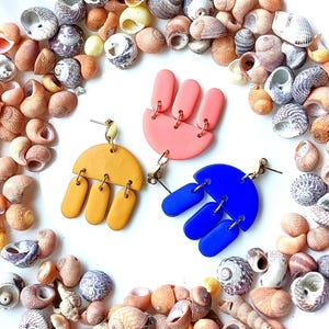 May include: Three pairs of colourful, handmade earrings. One pair is yellow, another is pink, and the third is blue. Each earring has a unique design with a half-circle top and dangling shapes. The earrings are displayed on a white surface surrounded by seashells.