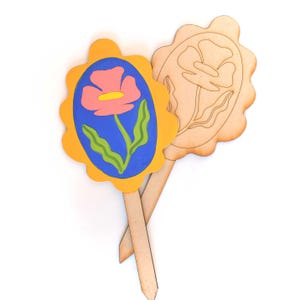 May include: Two wooden garden stakes. One is painted with a pink flower on a blue background, framed by a yellow border. The other is a natural wood cutout of a flower. Both have wooden stakes attached.
