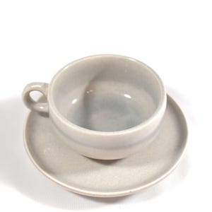 May include: A light gray ceramic teacup and saucer set. The cup has a rounded shape with a small handle. The saucer is round and flat. The set is made of a speckled ceramic material and is suitable for serving tea or coffee.