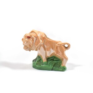 Może przedstawiać: Ceramiczna figurka lwa w odcieniach beżu i brązu, z czerwonymi akcentami w oczach. Lew stoi na zielonej podstawie, imitującej trawę. Figurka ma błyszczące wykończenie i szczegółowy, stylizowany design.