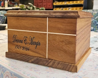 May include: A wooden urn, rectangular with a lid. The urn is a warm brown with a lighter wood inlay forming a cross. The name "Deanna E. Mangin" and the years "1941-2024" are engraved on the front, a memorial item.