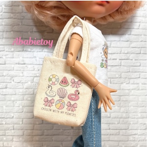 May include: A small, beige canvas tote bag featuring colorful summer-themed illustrations, including a watermelon slice, a flamingo, and a seashell. The text "CHILLIN WITH MY FLOATIES" is printed at the bottom. The bag has a simple handle and is held by a doll.