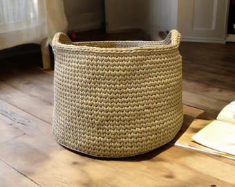 May include: A large, beige, crocheted storage basket with two handles. The basket is sitting on a wooden floor.