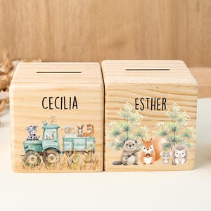 Può includere: Due salvadanai quadrati in legno chiaro con fessure per monete sulla parte superiore. La scatola a sinistra presenta un trattore verde con animali da fattoria e il nome CECILIA. Quella a destra mostra animali del bosco e il nome ESTHER.