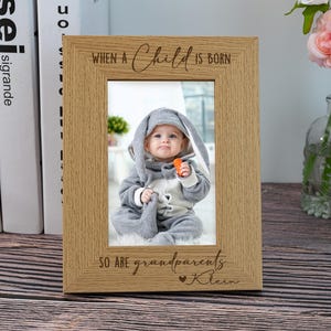 Puede incluir: Un marco de fotos de madera con el texto "Cuando nace un niño, también nacen los abuelos" y un corazón con el nombre "Klein" grabado en él. El marco tiene una foto de un bebé con un disfraz de conejito gris.