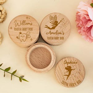 Op de afbeelding: Drie houten tandfee-doosjes met gepersonaliseerde namen en ontwerpen. De doosjes zijn rond en hebben een deksel. Eén doosje heeft een tand met een kroon en vleugels, een ander heeft een fee met vleugels en een toverstaf en de derde heeft een fee met vleugels.