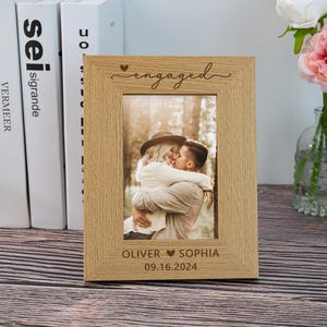 May include: A light-colored wooden picture frame with the word "engaged" and a heart etched at the top. The frame holds a photo of a couple embracing. The names "Oliver" and "Sophia" and the date "09.16.2024" are at the bottom.