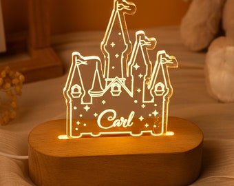 Puede incluir: Una luz nocturna de acrílico transparente con forma de castillo y brillo cálido, que presenta el nombre Carl grabado en letra cursiva y pequeños detalles de estrellas. La pieza de acrílico está montada sobre una base de madera ovalada.