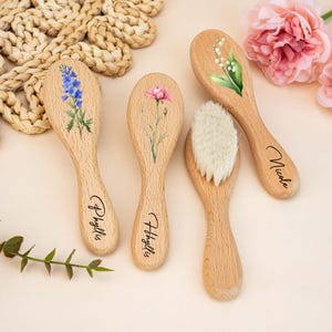 May include: Four wooden baby brushes with floral designs. One brush has a white bristle head. The brushes are decorated with blue, pink, and white flowers. The names "Phyllis" and "Nicole" are written on the handles.