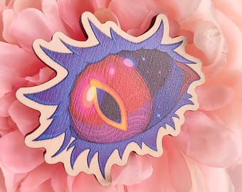 May include: Wooden pin featuring a stylized eye design with a vibrant color palette. The eye has a red and orange iris, surrounded by a blue and purple outer edge. The pin is set against a soft pink background. The website address zwaanyshop.etsy.com is visible.