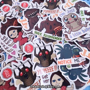 Peut inclure: Une collection d'autocollants colorés représentant divers personnages de dessins animés et des phrases. Les autocollants comprennent un monstre avec un panneau d'arrêt, une créature aux yeux rouges, une araignée et un personnage avec le texte "Good Boy!" et "100% Recommended".