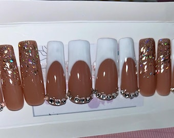 May include: Set of ten long, almond-shaped, fake nails with a white french tip design. The nails are a light brown color with gold glitter on the bottom half of the nails. Each nail has a row of small, clear rhinestones at the base.