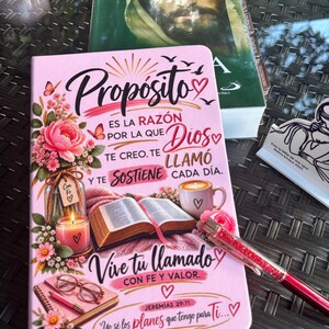 May include: A pink journal featuring floral illustrations, a candle, an open book, a coffee mug, and glasses. The text reads: Propósito es la razón por la que Dios te creó, te llamó y te sostiene cada día. Vive tu llamado con fe y valor. Jeremías 29:11. Yo sé los planes que tengo para ti.