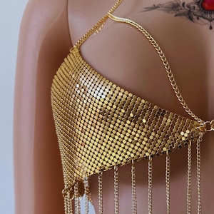 May include: A gold-colored chainmail top with a halter neck design. The top is made of small, circular metal pieces linked together, creating a shimmering effect. Delicate chains hang from the bottom edge, adding movement and detail to the garment.