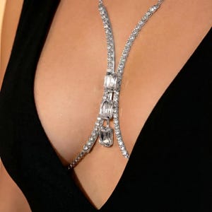 Peut inclure: Une cha&icirc;ne de corps argent&eacute;e orn&eacute;e de strass clairs et &eacute;tincelants. La cha&icirc;ne pr&eacute;sente un design en forme de Y avec trois pierres rectangulaires plus grandes comme point central. Le bijou est port&eacute; sur un v&ecirc;tement noir.