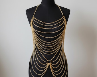 Puede incluir: Top de cota de malla de color dorado exhibido en un maniquí negro. El top estilo halter presenta múltiples hebras de delicadas cadenas que caen en cascada por la parte delantera, conectadas por un anillo circular en la cintura. Las cadenas están espaciadas uniformemente.