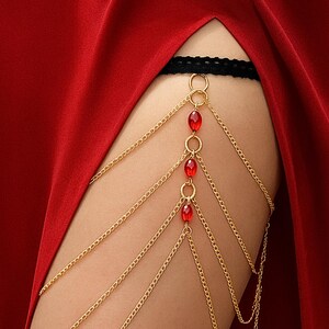 May include: Gold-colored body chain with red beads and a black lace band. The chain drapes over the thigh, with multiple strands and decorative red beads. The background is a solid red fabric.
