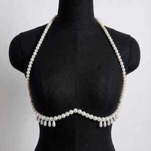 K&ouml;nnte beinhalten: Eine wei&szlig;e Perlen-K&ouml;rperkette. Die Kette ist &uuml;ber einer schwarzen Schaufensterpuppe drapiert. Die Kette hat einen Neckholder-Stil mit einer Reihe kleiner Perlen und einem Fransenbehang aus tropfenf&ouml;rmigen Perlen.