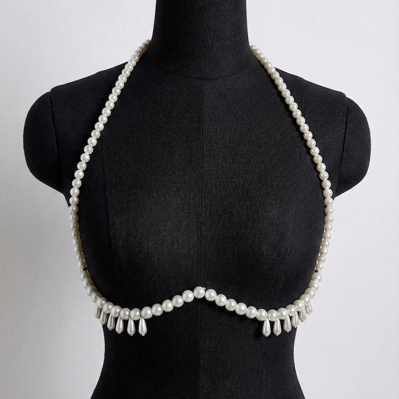 K&ouml;nnte beinhalten: Eine wei&szlig;e Perlen-K&ouml;rperkette. Die Kette ist &uuml;ber einer schwarzen Schaufensterpuppe drapiert. Die Kette hat einen Neckholder-Stil mit einer Reihe kleiner Perlen und einem Fransenbehang aus tropfenf&ouml;rmigen Perlen.