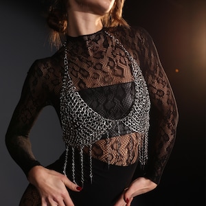 May include: A silver chainmail top with dangling chain fringe. The top is worn over a black lace long-sleeve top and black garment. The image is set against a dark background.