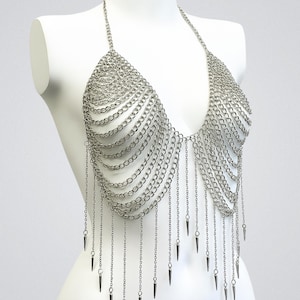 May include: A silver-toned metal chainmail halter top featuring multiple draped layers of interlocking circular links. The garment includes a neck strap and a series of vertical chain fringes that terminate in small, pointed metal spikes at the bottom.