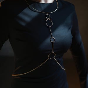 K&ouml;nnte beinhalten: Eine goldfarbene K&ouml;rperkette mit minimalistischem Design. Die Halskette hat einen oberen Teil im Choker-Stil, eine Reihe von verbundenen Kreisen und Linien sowie eine Kette, die &uuml;ber den Oberk&ouml;rper drapiert ist. Der Schmuck wird &uuml;ber einem schwarzen Kleid getragen.