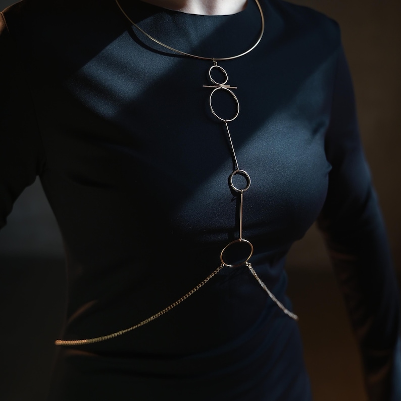 K&ouml;nnte beinhalten: Eine goldfarbene K&ouml;rperkette mit minimalistischem Design. Die Halskette hat einen oberen Teil im Choker-Stil, eine Reihe von verbundenen Kreisen und Linien sowie eine Kette, die &uuml;ber den Oberk&ouml;rper drapiert ist. Der Schmuck wird &uuml;ber einem schwarzen Kleid getragen.