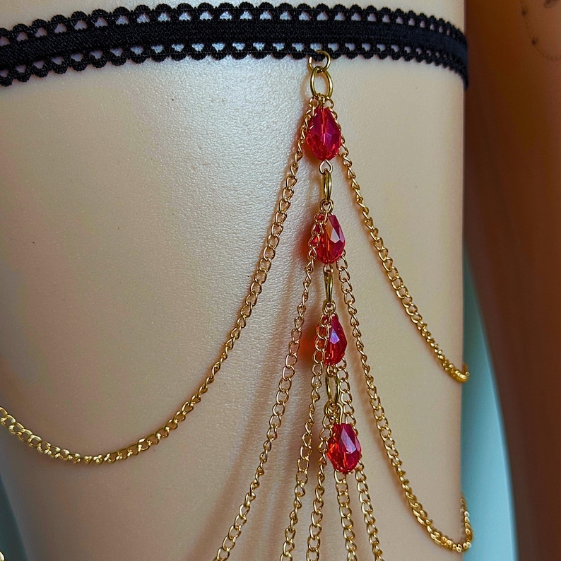 May include: A black lace garter with gold-colored chains and red teardrop-shaped beads. The chains hang down from the garter, creating a layered effect. The garter is worn on the thigh.
