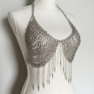 May include: A silver-toned chainmail halter top constructed from interlocking metal rings. The garment features a draped, scalloped neckline and a series of dangling chain fringe accents, each finished with a small, pointed metal spike at the end.